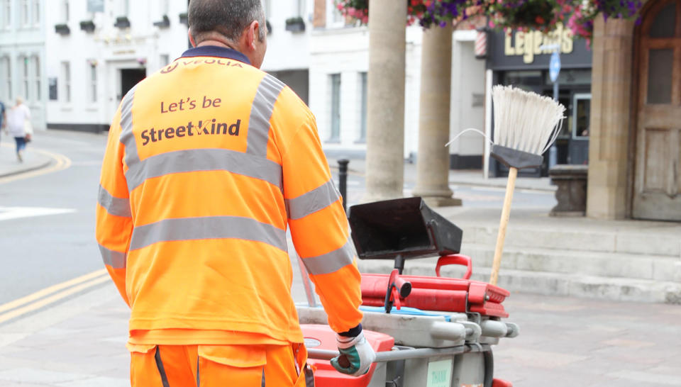 A sweeper walks away from the camera pushing a sweeper's cart. 'Let's be StreetKind' is printed on the back of his uniform.