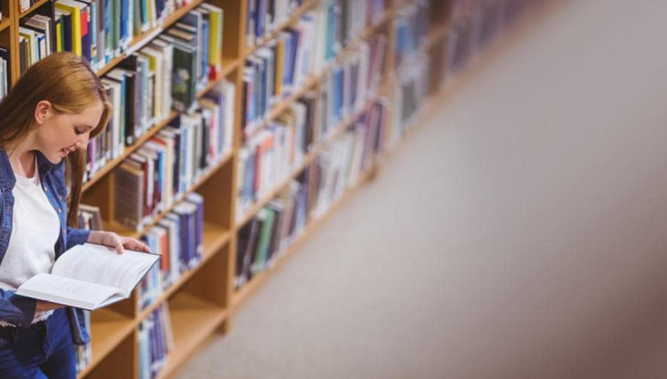 woman leaning on book shelf in a libairy reading a book 