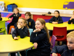 Primary school pupils sit in a colourful classroom with two pupils gazing at an intriguing presentation happening off screen.