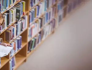 woman leaning on book shelf in a libairy reading a book 