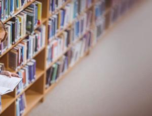 woman leaning on book shelf in a libairy reading a book 