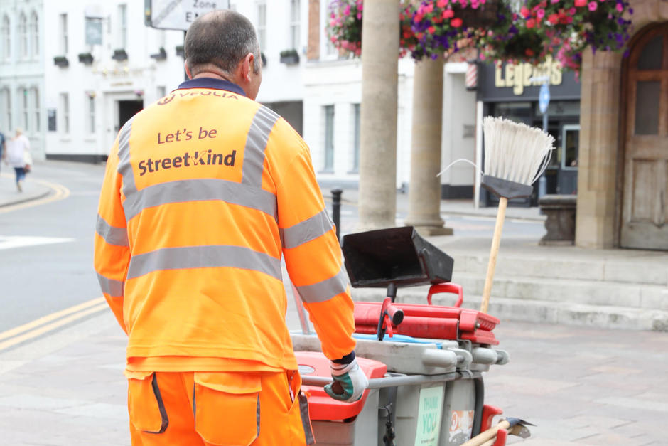 A sweeper walks away from the camera pushing a sweeper's cart. 'Let's be StreetKind' is printed on the back of his uniform.