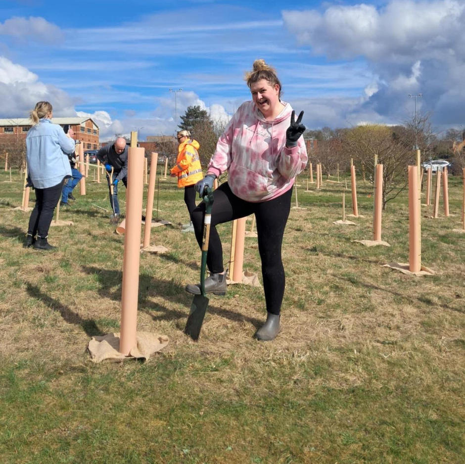 A member of the Customer Excellence Team smiles at the camera with her foot on a spade ready to dig a hole for a sapling.