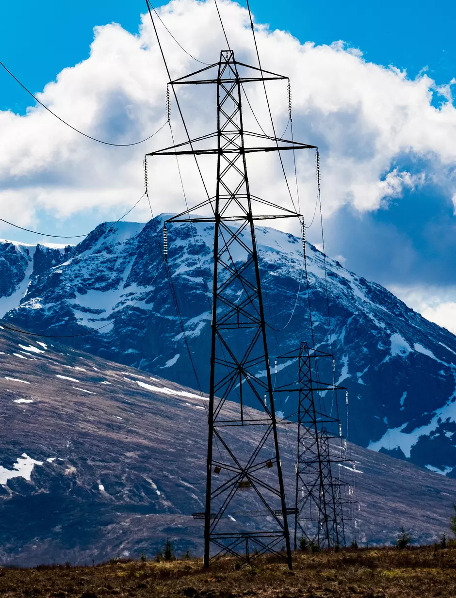 An electricity pylon rises in front of a snow capped mountain with blue sky and white clouds in the background.