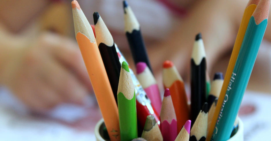A mixture of colouring pencils of different lengths and colours in a pot, with a child in the background colouring