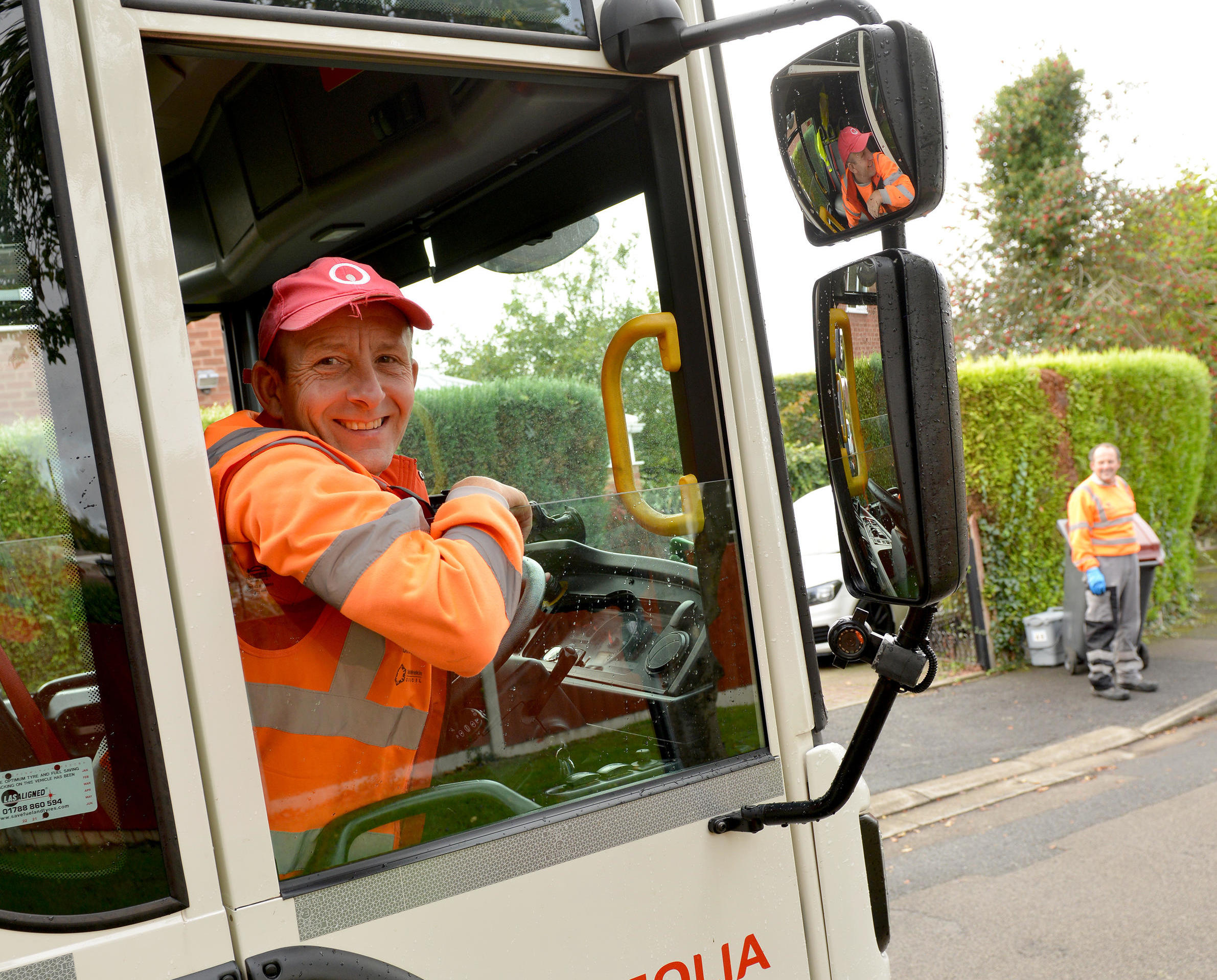 Photo showing a cheerful driver in a bin collection truck, with another crew member in the backgroun collecting bins.